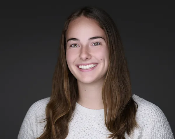 Headshot of a young woman with long brown hair in a white top on a dark gray background